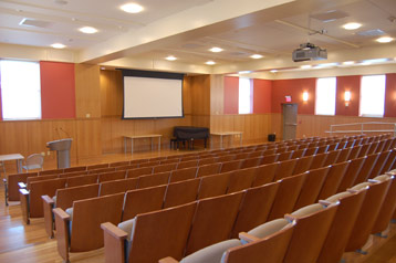A large auditorium with rows of chairs that are bolted to the floor. In front of the room is a podium and a pulled down projector screen.