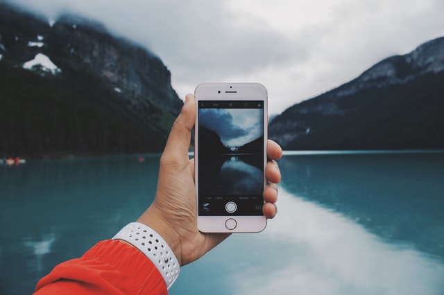 A hand holding up an iPhone in front of a beautiful mountain lake.