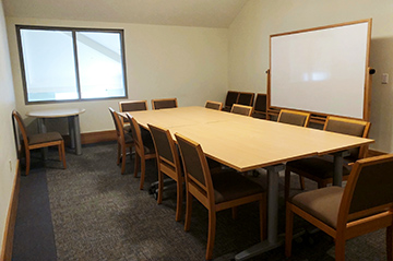 Large table to the right surrounded by chairs with a whiteboard behind the table. On the left is a window and a second smaller table beneath it.