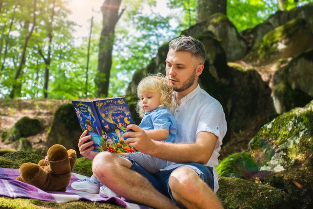 Father holds child on lap reading a book while sitting outside.
