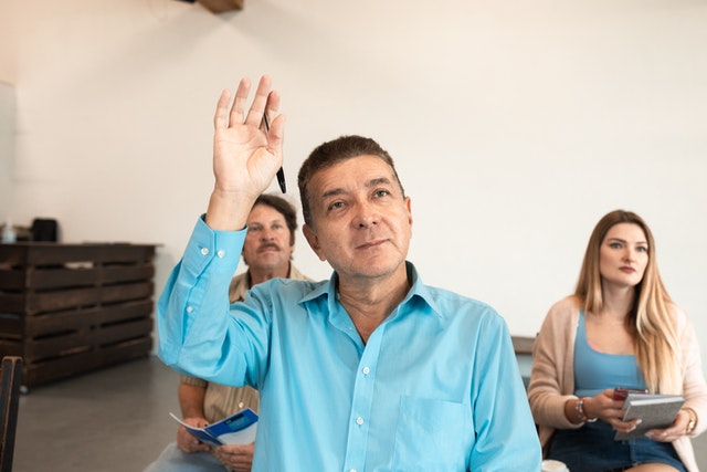 Man sitting in classroom holding up his hand. Two other students are visible behind him.