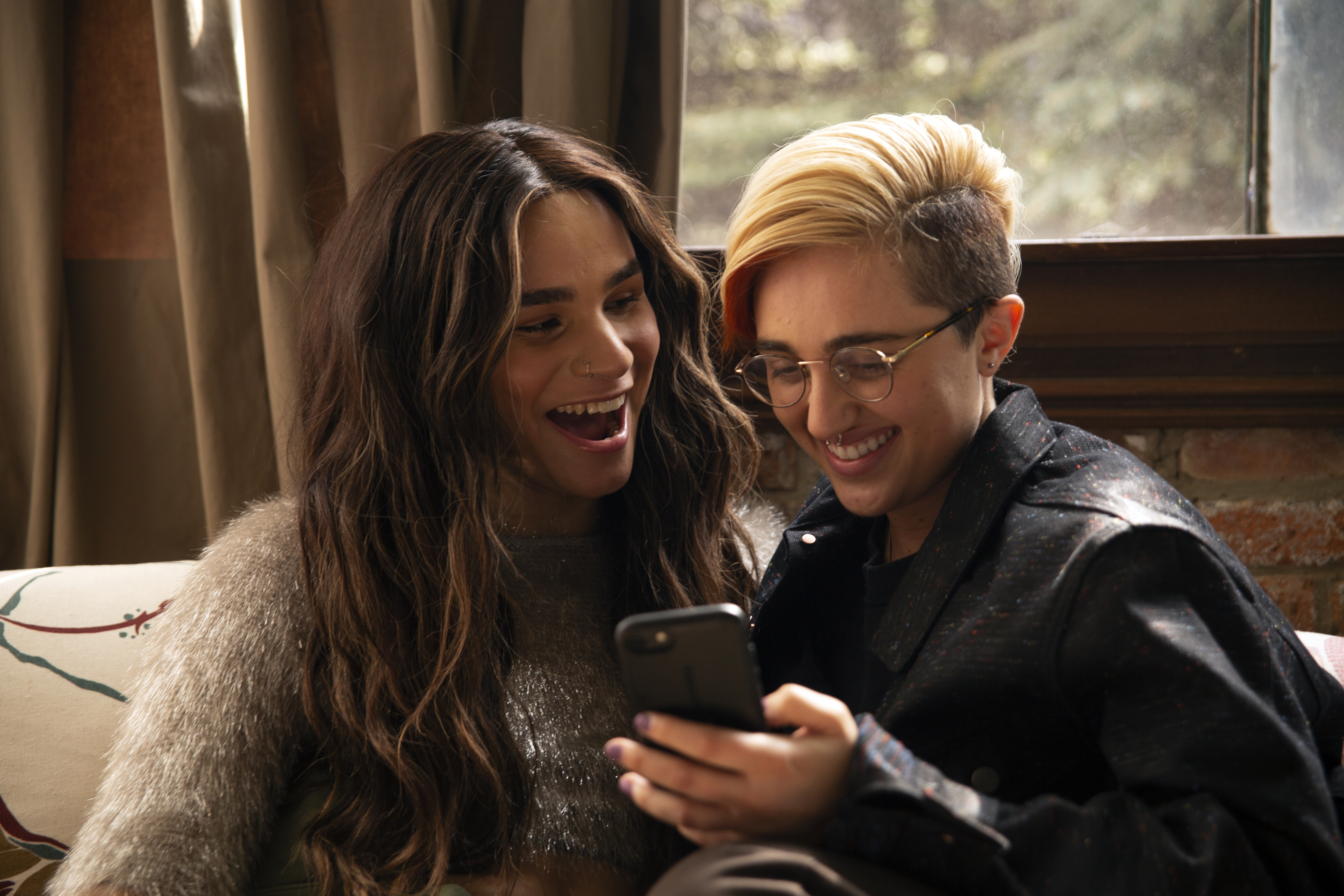 A transfeminine non-binary person and transmasculine gender-nonconforming person looking at a phone and laughing
