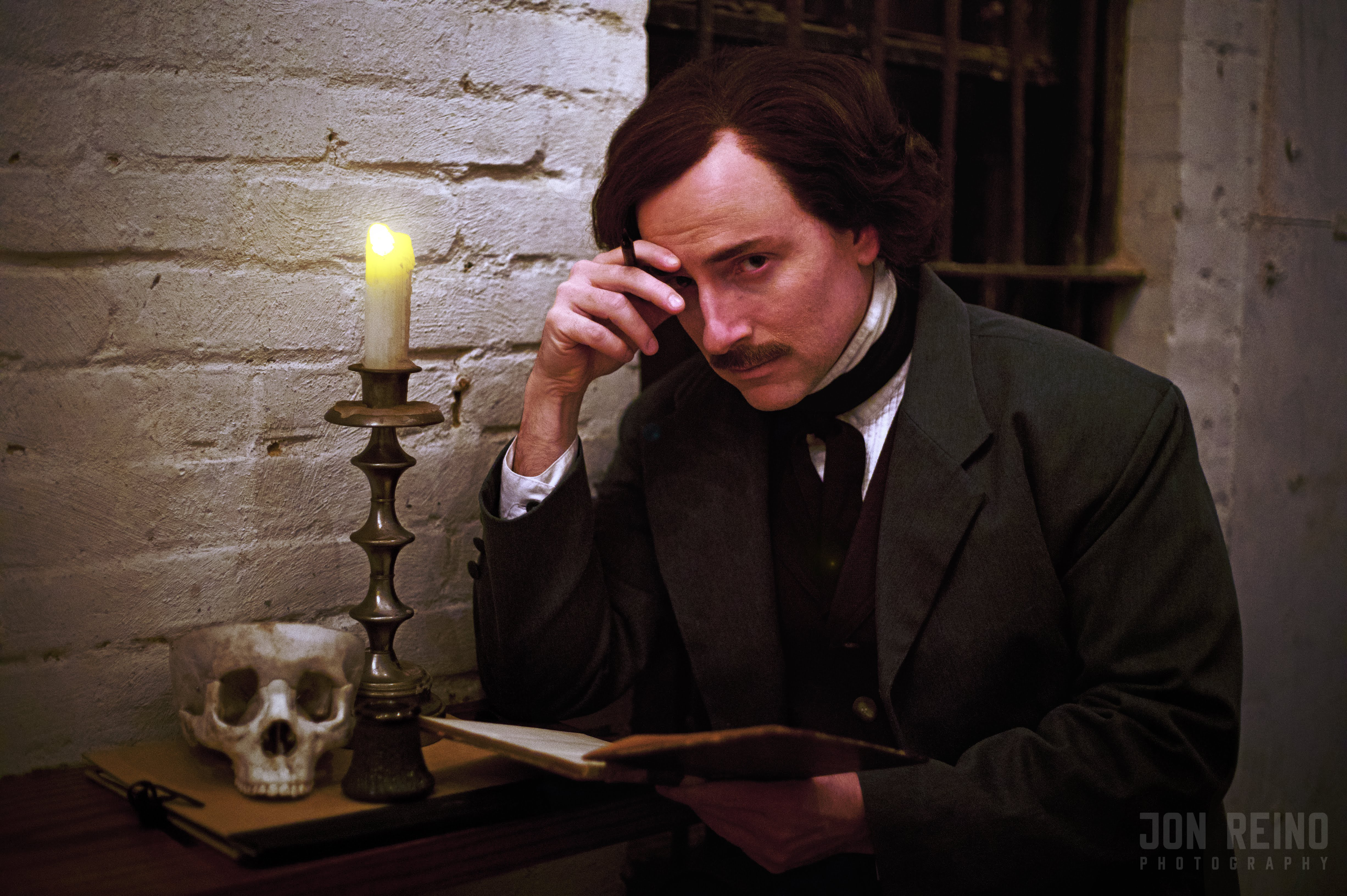 A man resembling Edgar Allan Poe sits at desk near lit candle