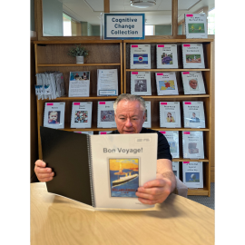 A white man with gray hair and a black t-shirt reads a book titled "Bon Voyage!" The book is part of our Cognitive Change collection, which is shown in the background.