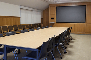 A large table dominates the left side of the image with chairs around the table. At the back of the room is a large built-in screen.