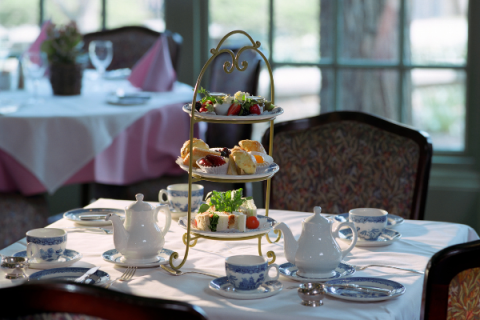 Table covered in white tablecloth, three tiered stand housing fruit, desserts, and tea sandwiches sits on center of table, surrounded by tea pot and blue and white tea cups. 