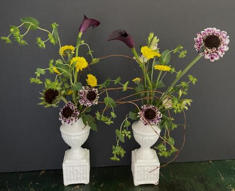 image of two white urns with an assortment of late summer flowers in both, like sunflowers and a type of lily