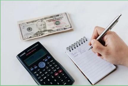 image of a calculator, money and a note pad and a hand with a pen writing something probably a number.